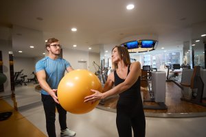 personal trainer: Female and male are holding a yellow fitball in their hands, they are in a modern gym